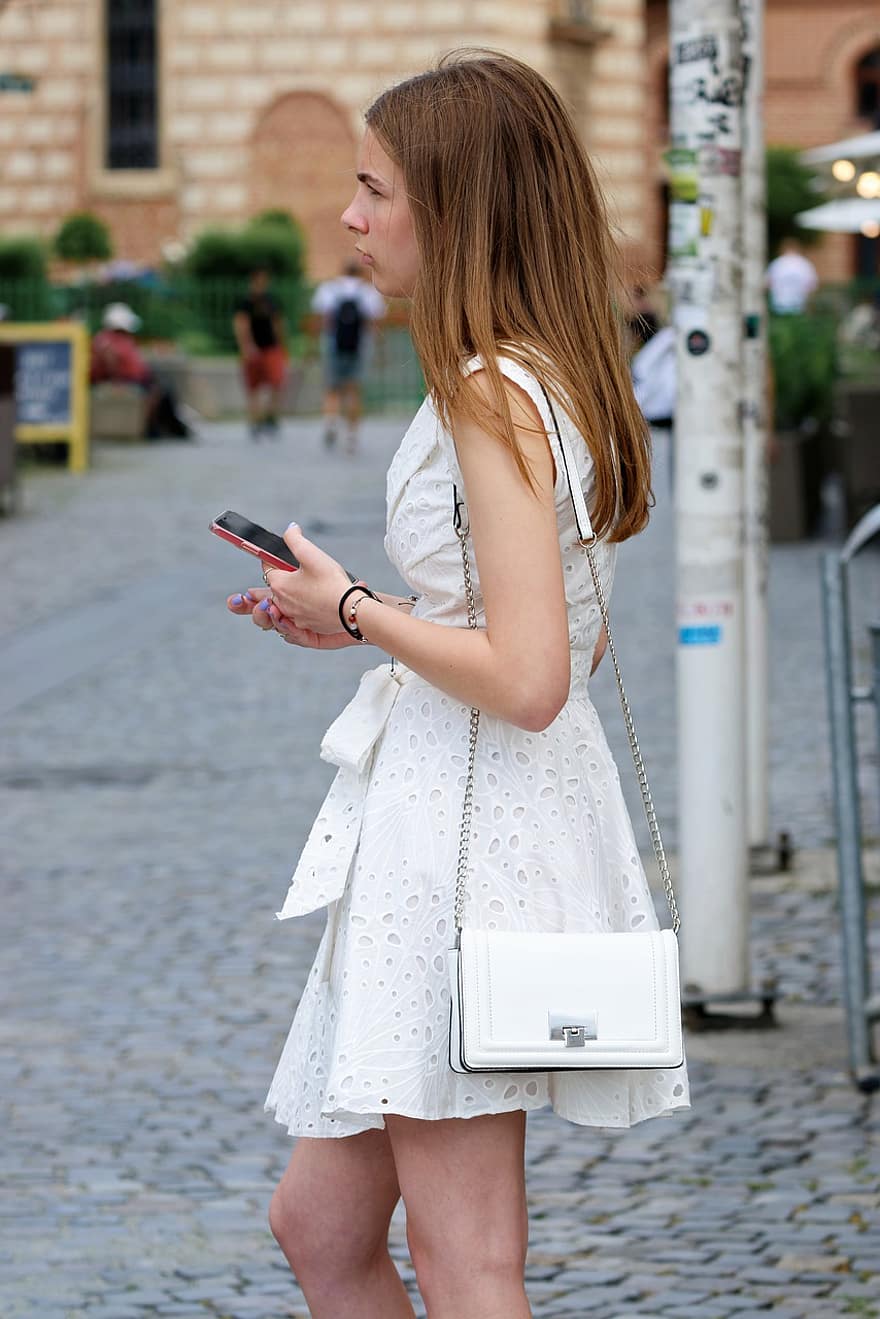 niña, bonita, vestido blanco, bolso, teléfono inteligente, en pie, esperando, pavimento, calle, urbano, una persona